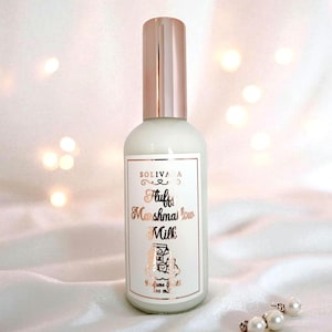 Puede incluir: Un frasco de perfume blanco con tapa y etiqueta de oro rosa. La etiqueta dice "Fluffy Marshmallow Milk" en una fuente decorativa. La botella está sobre una superficie de satén blanco con un collar de perlas.