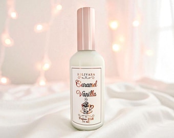 May include: A white Solivara Caramel Vanilla perfume bottle with a pink cap. The label displays the scent name in script, a coffee cup illustration, and "Perfume & Milk 100 mL". The background is soft and out of focus, with a hint of fairy lights.