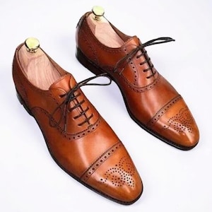 Könnte beinhalten: Ein Paar hellbraune Oxford-Schuhe aus Leder mit einer Zehenkappe mit dekorativen Brogue-Perforationen, dunkelbraunen Schnürsenkeln und hellen Holz-Schuhspannern in den Fersen. Die Schuhe haben eine polierte Oberfläche und einen flachen Absatz.
