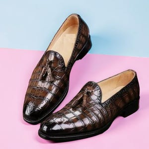 May include: A pair of brown loafers featuring a glossy crocodile-embossed texture, decorative tassels on the vamp, and intricate brogue detailing along the edges. The shoes have a low heel, a rounded toe, and a light-coloured interior lining.