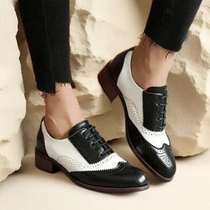 May include: Two-tone Oxford shoes in black and white with a low heel. The shoes have a traditional wingtip design with decorative perforations. The uppers are a mix of black and white leather, with a brown heel and sole.