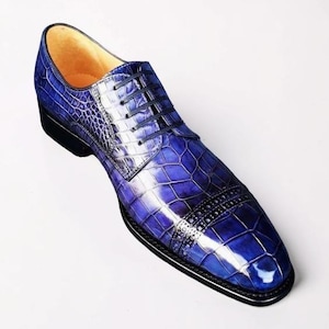 Könnte beinhalten: Ein glänzender blauer Oxford-Schuh mit texturiertem Krokodilleder-Muster, einer Zehenkappe mit dekorativen Brogue-Perforationen, dünnen schwarzen Schnürsenkeln und einer soliden schwarzen Sohle. Das Innenfutter des Schuhs ist hellbeige.