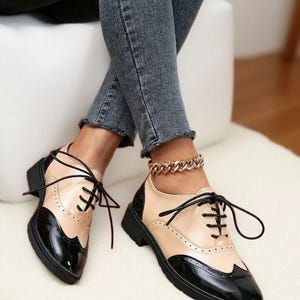 May include: Two-tone oxford shoes in black and beige leather. The shoes feature a classic wingtip design, black laces, and a low heel. The shoes are worn with denim jeans.
