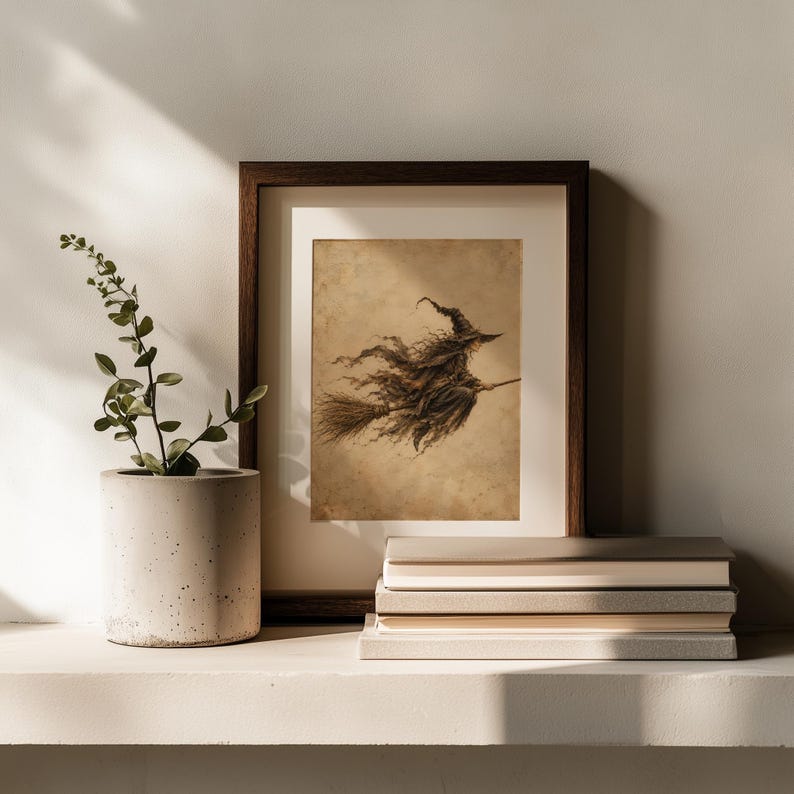 May include: A framed art print featuring a sepia-toned illustration of a witch flying on a broomstick. The artwork is displayed in a dark brown wooden frame, positioned next to a light gray cylindrical planter with a green plant and a stack of books.