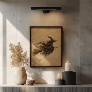 May include: A framed wall art print featuring a sepia-toned illustration of a witch flying on a broomstick. The artwork is displayed in a simple black frame, positioned on a wall with a light fixture above and decorative items below.