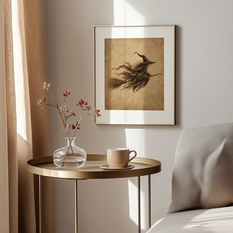 May include: A framed wall art print featuring a sepia-toned illustration of a witch flying on a broomstick. The artwork is displayed in a thin gold frame, hanging on a light-colored wall above a round gold side table with a clear glass vase.