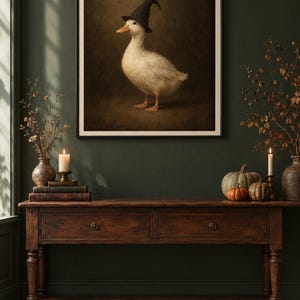 K&ouml;nnte beinhalten: Ein gerahmter Wandkunstdruck, der eine wei&szlig;e Ente mit einem schwarzen, spitzen Hexenhut zeigt. Die Ente ist im Profil vor einem dunklen, stimmungsvollen und strukturierten braunen Hintergrund dargestellt. Das Bild ist in einem schmalen schwarzen Rahmen.