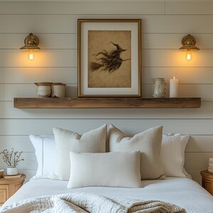 May include: A bedroom scene featuring a wooden floating shelf mounted on a white shiplap wall. The shelf holds a framed illustration of a witch on a broomstick, two ceramic jars, a small pitcher, and a white pillar candle, flanked by two brass wall lamps.