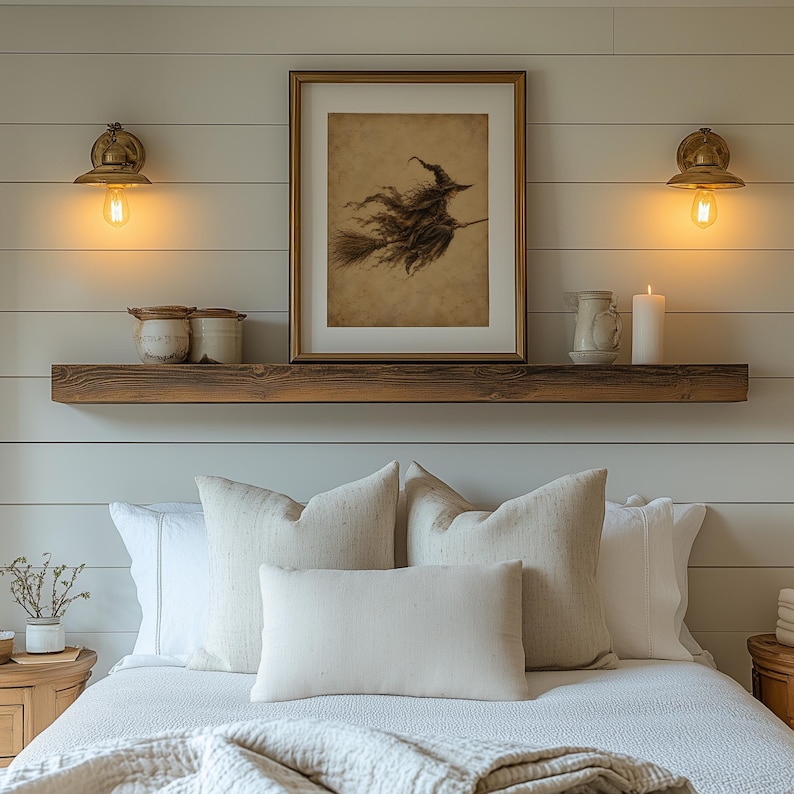 May include: A bedroom scene featuring a wooden floating shelf mounted on a white shiplap wall. The shelf holds a framed illustration of a witch on a broomstick, two ceramic jars, a small pitcher, and a white pillar candle, flanked by two brass wall lamps.