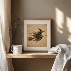 May include: A framed wall art print featuring a sepia-toned illustration of a figure in a pointed hat riding a broomstick. The artwork is displayed in a light wood frame, positioned next to a white ceramic mug and a small vase with dried branches.
