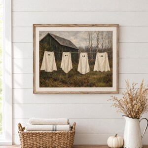 May include: A framed wall art print depicting a rustic wooden barn in a field with four white ghost-shaped sheets hanging on a clothesline. The scene features muted earth tones, bare trees, and a cloudy sky, capturing a seasonal autumn atmosphere.
