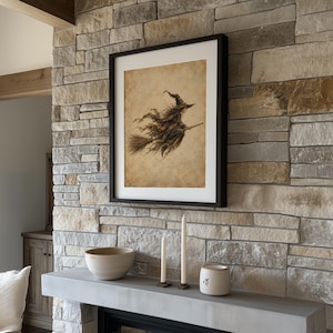 May include: A framed wall art print featuring a sepia-toned illustration of a witch flying on a broomstick. The artwork is displayed in a simple black frame with a white mat, hanging on a textured stone wall above a mantel with two candles.