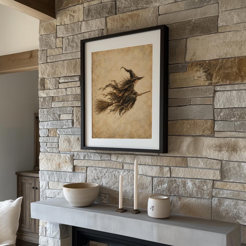 May include: A framed wall art print featuring a sepia-toned illustration of a witch flying on a broomstick. The artwork is displayed in a simple black frame with a white mat, hanging on a textured stone wall above a mantel with two candles.