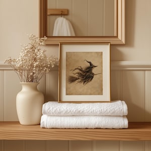 May include: A framed print featuring a sepia-toned illustration of a witch flying on a broomstick, displayed next to a matte cream ceramic vase filled with dried baby's breath flowers, resting upon two neatly folded white textured towels.