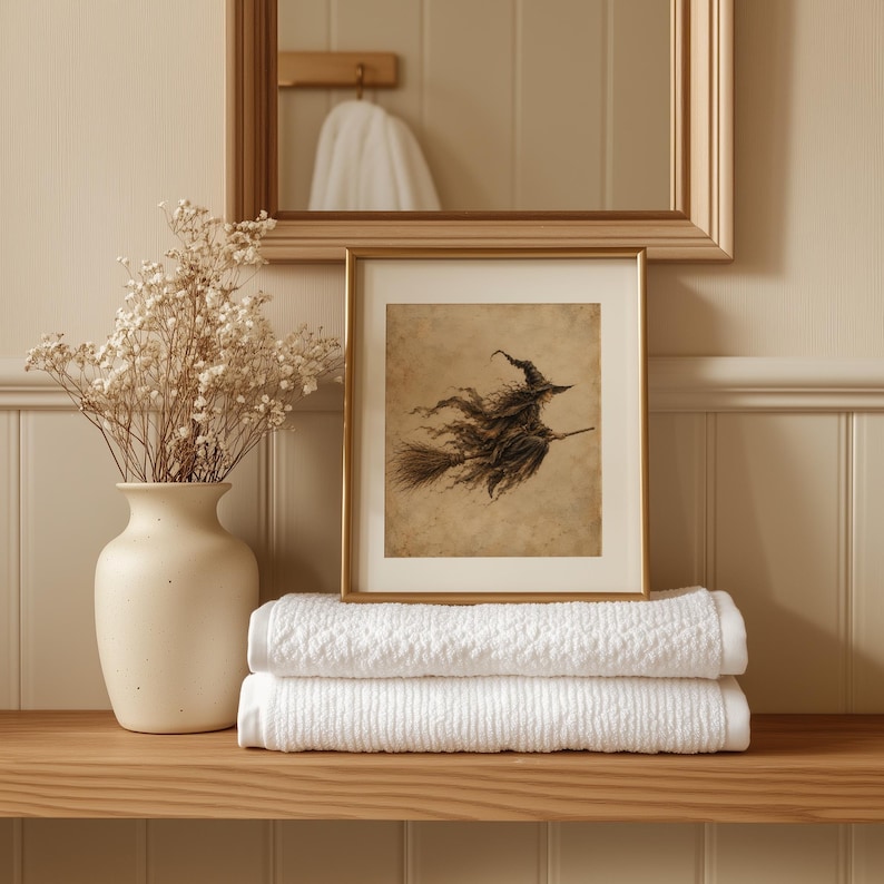 May include: A framed print featuring a sepia-toned illustration of a witch flying on a broomstick, displayed next to a matte cream ceramic vase filled with dried baby's breath flowers, resting upon two neatly folded white textured towels.