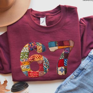 Könnte beinhalten: Ein weinrotes Sweatshirt mit der Zahl "67" auf der Vorderseite in einem Patchwork-Design. Das Bild enthält eine blaue Jeans, einen braunen Hut und eine Kaffeetasse. Das Wort "MAROON" ist in der oberen rechten Ecke aufgedruckt.