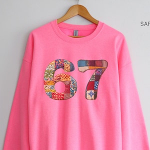 May include: A bright pink sweatshirt with the number "67" appliqued on the front. The numbers are made of a patchwork of colorful fabric squares. The text "SAFETY PINK" is in the upper right corner.