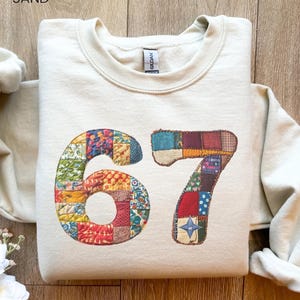 Könnte beinhalten: Ein sandfarbener Pullover mit der Zahl "67" auf der Vorderseite. Die Zahlen bestehen aus buntem Patchwork-Stoff. Das Wort "SAND" ist oben im Bild aufgedruckt.