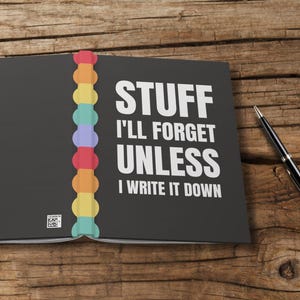 Puede incluir: Cuaderno abierto con el texto "STUFF I'LL FORGET UNLESS I WRITE IT DOWN" en blanco. El cuaderno tiene una cubierta negra y un lomo colorido. Un bolígrafo negro descansa sobre una superficie de madera.