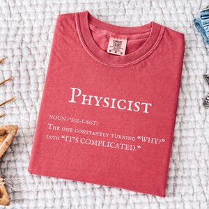 May include: Crimson-colored t-shirt with the word "Physicist" printed in white. The shirt includes a definition and the phrase "The one constantly turning 'why?' into 'it's complicated.'" The shirt is folded on a white textured surface.