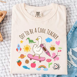 Peut inclure: T-shirt ivoire avec l'inscription "Off to be a Cool Teacher" et un dessin d'un canard sur un skateboard tenant un crayon, entouré d'illustrations colorées. Le t-shirt est posé sur une surface blanche à côté d'un jean bleu.
