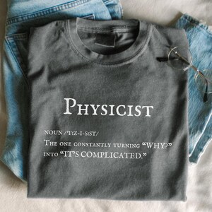 May include: A gray t-shirt with the word "PHYSICIST" printed in white, along with a definition. The shirt is paired with light blue jeans and eyeglasses. The text on the shirt reads: "The one constantly turning 'WHY?' into 'IT'S COMPLICATED.'"