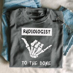 Könnte beinhalten: Ein graues T-Shirt mit dem Wort "RADIOLOGIST" in einem weißen Rechteck und einer weißen Skeletthand, die eine "Hang Loose"-Geste macht. Der Text "TO THE BONE" befindet sich unter der Hand. Hellblaue Jeans und eine Brille sind ebenfalls im Bild.