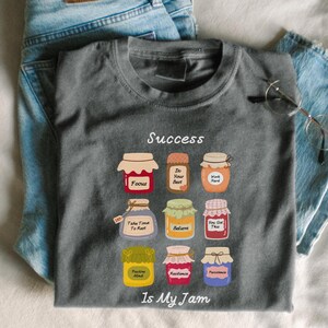 May include: A gray t-shirt with the word "Success" above a grid of illustrated jam jars, each labeled with motivational words like "Focus" and "Believe." The phrase "Is My Jam" is printed below the jars. Light blue jeans are in the background.
