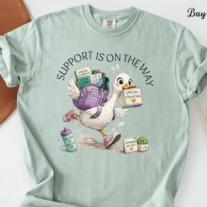 Könnte beinhalten: Ein hellgrünes T-Shirt mit einer Grafik einer weißen Gans, die einen lila Rucksack, eine Wasserflasche und Blöcke mit der Aufschrift "Special Education" und "Different Not Less" trägt. Der Text "Support Is On The Way" steht über der Gans.