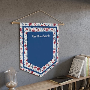 May include: A pennant-shaped wall hanging with a blue centre and floral patterned border in red and white. The text "Use It or Lose It" is displayed in white script. The pennant is suspended from a wooden dowel.