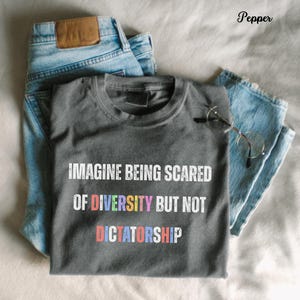 May include: A gray t-shirt with the text "IMAGINE BEING SCARED OF DIVERSITY BUT NOT DICTATORSHIP" in white and colorful letters. The shirt is laid on top of a pair of light blue jeans.