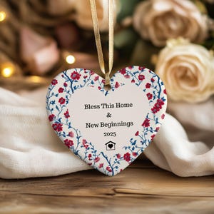 May include: Heart-shaped ceramic ornament with a floral pattern in red and blue on a white base. The ornament features the text "Bless This Home & New Beginnings 2025" and a house icon. A gold ribbon is attached for hanging.