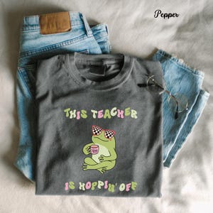 Könnte beinhalten: Ein graues T-Shirt mit dem Text "THIS TEACHER IS HOPPIN' OFF" und einem Cartoon-Frosch mit Sonnenbrille und einer Tasse. Das Shirt liegt gefaltet neben einer Jeans und einer Brille.