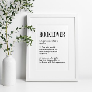 May include: A framed print with the word "BOOKLOVER" in bold black letters. Below, three definitions of a booklover are listed in black text. A white vase with greenery is to the left.