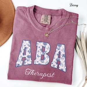 Op de afbeelding: Bessenkleurig T-shirt met de letters "ABA" in een bloemenpatroon en het woord "Therapist" eronder. Een gouden ketting met een hanger en een bruine hoed zijn ook zichtbaar.