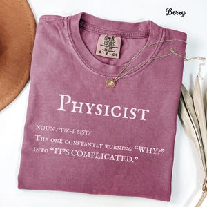 May include: Berry-colored t-shirt with the word "PHYSICIST" in white block letters. The shirt also includes the definition of physicist and the phrase "The one constantly turning 'WHY?' into 'IT'S COMPLICATED.'" A gold necklace with a pendant rests on the shirt.