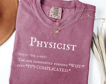 May include: A berry-colored short-sleeve t-shirt with a crew neckline. The shirt features white text that reads: PHYSICIST NOUN /'FIZ-I-SIST/ The one constantly turning "WHY?" into "IT'S COMPLICATED." A thin gold-toned necklace rests on the shirt.
