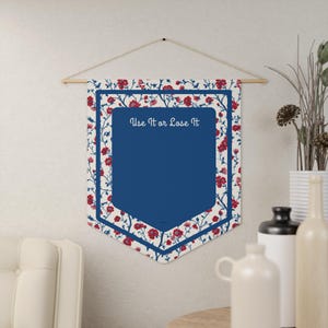 May include: A wall hanging banner with a blue central panel and a floral border in red and white. The banner is suspended from a wooden rod and features the text "Use It or Lose It".
