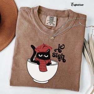 May include: An espresso-colored crew neck t-shirt featuring a graphic of a black cat wearing a red beret and scarf, sitting inside a white bowl. The text "le Sigh" is printed in black next to the cat. The shirt has a Comfort Colors brand label.