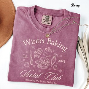 Può includere: T-shirt color bacca con la scritta "Winter Baking Social Club" e grafica a tema panificazione. La maglietta ha girocollo e maniche corte. Una collana d'oro con un ciondolo è drappeggiata sulla maglietta.