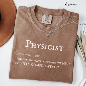 May include: Espresso-colored t-shirt with the word "PHYSICIST" in white block letters. The shirt includes a definition and quote about physicists. A silver necklace with a pendant rests on the shirt.