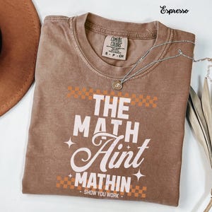 Op de afbeelding: Espresso-kleurig T-shirt met de witte tekst "THE MATH Aint MATHIN". Het shirt heeft een ronde hals en korte mouwen. Een zilveren ketting met een hanger is over het shirt gedrapeerd. Het shirt is van het merk Comfort Colors.