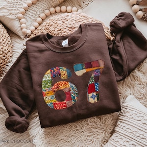 Könnte beinhalten: Dunkelbraunes Sweatshirt mit der Zahl "67" auf der Vorderseite. Die Zahlen bestehen aus buntem Patchwork-Stoff. Das Sweatshirt hat einen Rundhalsausschnitt und lange Ärmel. Der Text "DARK CHOCOLATE" steht unten.
