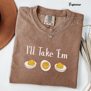 May include: An espresso-colored short-sleeve t-shirt with a crew neckline. The shirt features the white text 'I'll Take 'Em' above three illustrations of halved hard-boiled eggs. A silver-toned necklace with a small circular pendant rests on the shirt.