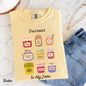 May include: Pale yellow t-shirt featuring the phrase "Success Is My Jam" and illustrated jam jars with motivational labels such as "Focus" and "Believe." The shirt has a round neck and short sleeves.