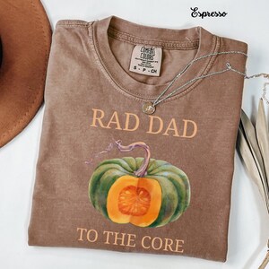 May include: Espresso-coloured t-shirt featuring the text "RAD DAD TO THE CORE" and a watercolour-style pumpkin graphic. The shirt has a crew neck and short sleeves. A silver necklace with a pendant is draped across the shirt.