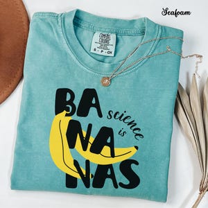 May include: A seafoam green t-shirt featuring a graphic of a yellow banana and the black text "BANANAS" with the word "science" and "is" in smaller black script. The shirt has a Comfort Colors brand label at the neck indicating size small.