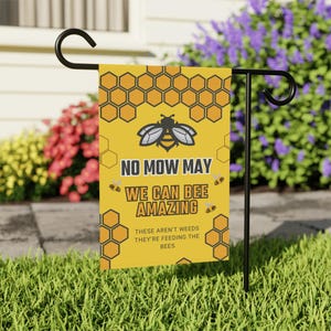 May include: A yellow garden flag with a bee and honeycomb design. The flag reads "NO MOW MAY, WE CAN BEE AMAZING, THESE AREN'T WEEDS THEY'RE FEEDING THE BEES." The flag is displayed on a black metal stand in a garden.