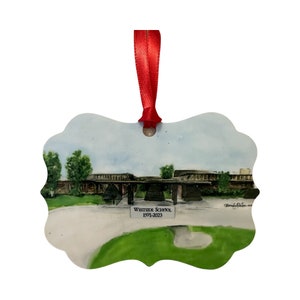 May include: Ornament featuring a watercolor painting of Westside School. The image shows the school building, green grass, and trees under a cloudy sky. The text "Westside School 1971-2023" is visible on the ornament, which has a red ribbon for hanging.