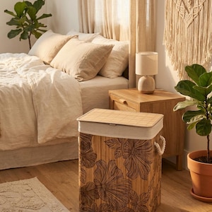 May include: A rectangular bamboo laundry hamper with a floral design. The hamper has a light-colored fabric liner and a bamboo lid. The floral design is a dark brown color. The hamper is in a bedroom setting.